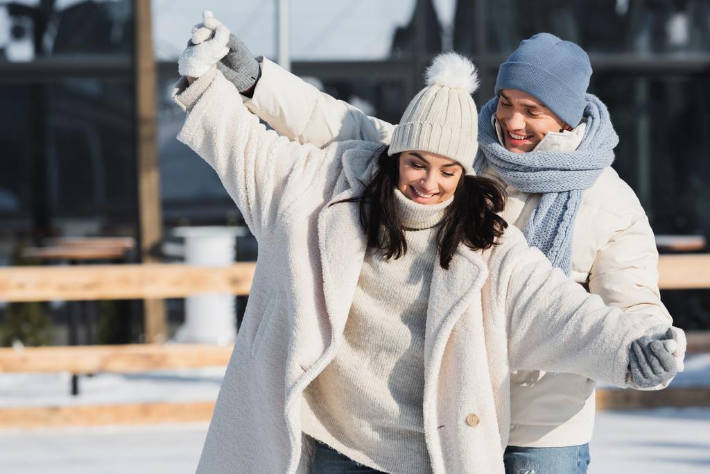 счастливый молодой человек в зимней шляпе, держа за руки улыбающуюся девушку с протянутыми руками  - Фото, изображение