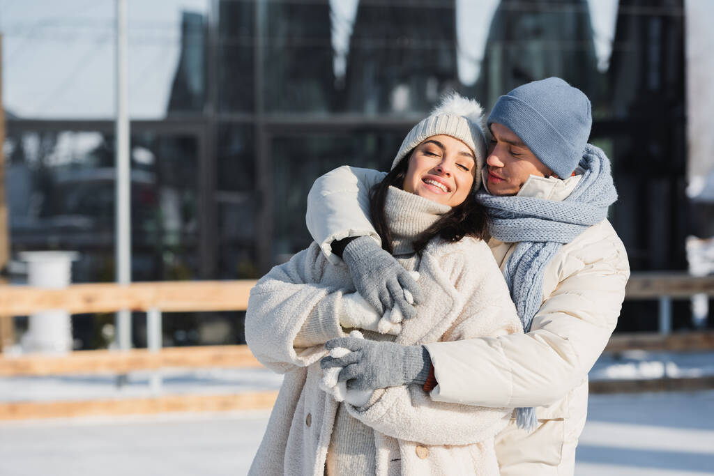 feliz joven hombre y mujer en sombreros de invierno abrazándose en pista de hielo - Foto, Imagen