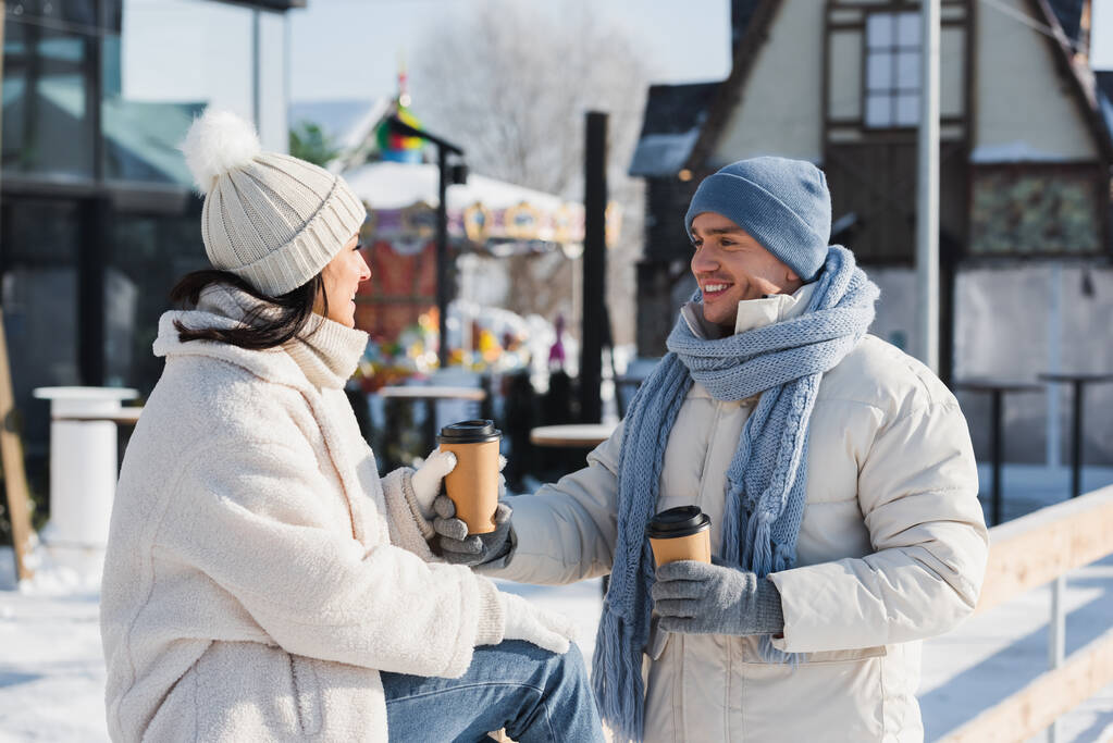 χαρωπός άνθρωπος δίνει χάρτινο κύπελλο στην ευτυχισμένη φίλη του στο χειμερινό καπέλο - Φωτογραφία, εικόνα