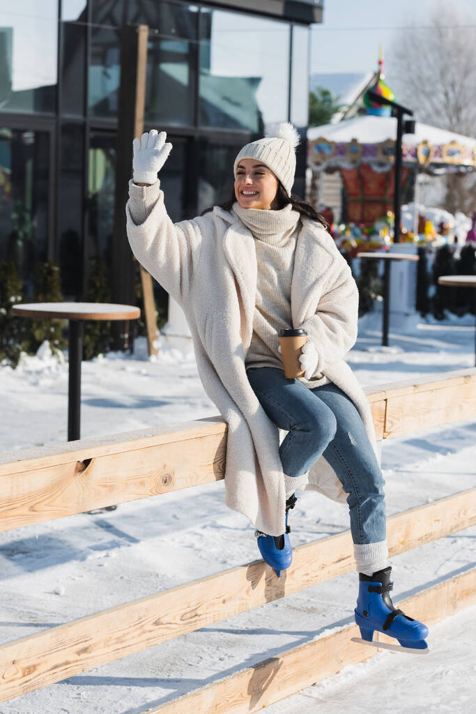 longitud completa de la joven feliz en el sombrero de invierno y patines de hielo sosteniendo taza de papel y agitando la mano cerca de pista de hielo - Foto, imagen