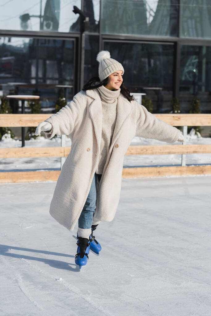volledige lengte van gelukkig jong vrouw in winter hoed en jas schaatsen op ijsbaan - Foto, afbeelding