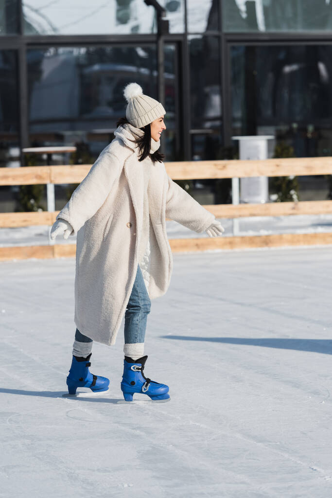 volledige lengte van vrolijke jonge vrouw in winter hoed en jas schaatsen op ijsbaan - Foto, afbeelding