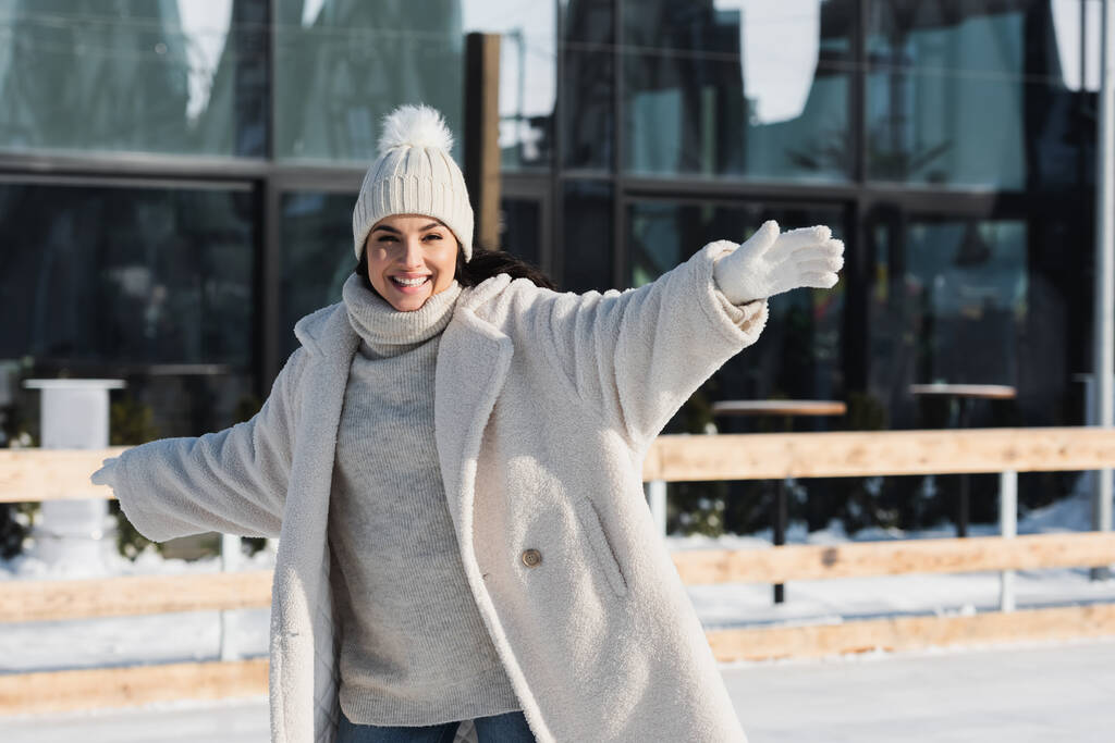 θετική νεαρή γυναίκα σε χειμερινό καπέλο και παλτό πατινάζ σε παγοδρόμιο - Φωτογραφία, εικόνα