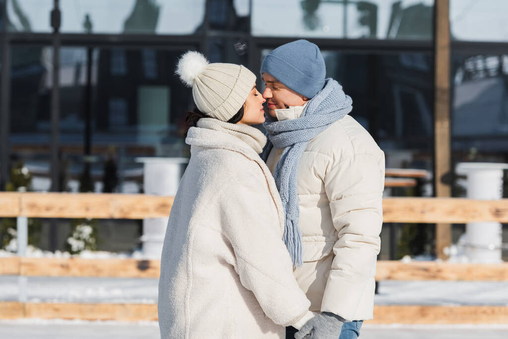 młoda szczęśliwa para w czapkach zimowych trzymając się za ręce i całując na zewnątrz  - Zdjęcie, obraz
