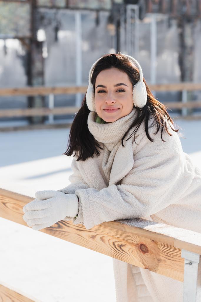 feliz jovem mulher no ouvido muffs inclinando-se na borda de madeira na pista de gelo  - Foto, Imagem