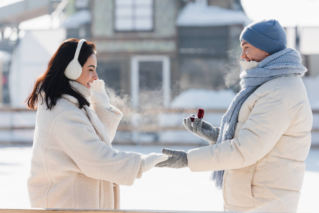 vista laterale dell'uomo in inverno cappello scatola di tenuta con anello di nozze vicino felice giovane donna in manicotti orecchio - Foto, immagini