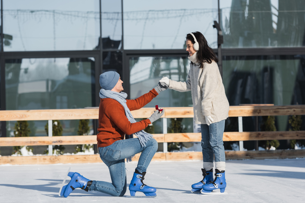 man holding box met trouwring en knielen terwijl het maken van aanzoek aan gelukkige vrouw in oor moffen op de ijsbaan - Foto, afbeelding