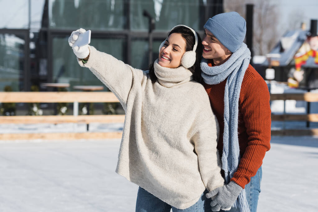 Ευτυχισμένη γυναίκα με πουλόβερ και ωτοασπίδες που βγάζει σέλφι με τον φίλο της σε παγοδρόμιο.  - Φωτογραφία, εικόνα