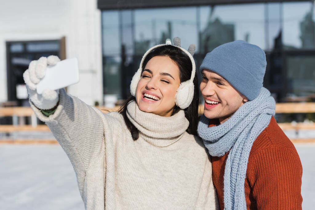felice donna in maglione e paraorecchie prendendo selfie con allegro fidanzato sulla pista di pattinaggio  - Foto, immagini