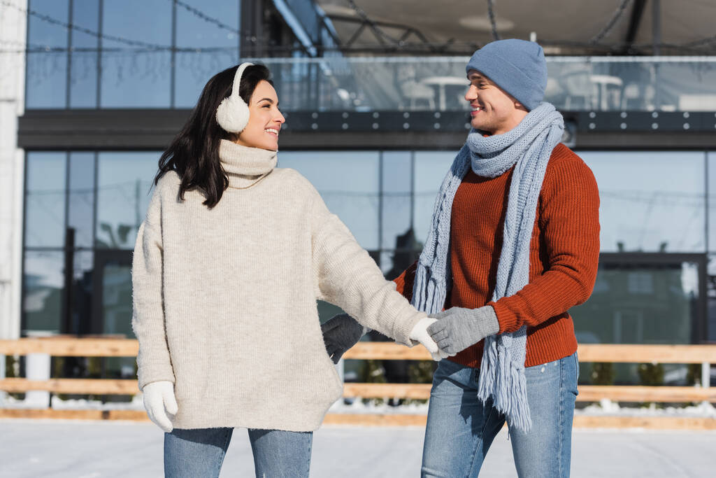 glückliche junge Frau im Ohrenschutz hält Händchen mit fröhlichem Freund auf der Eisbahn  - Foto, Bild