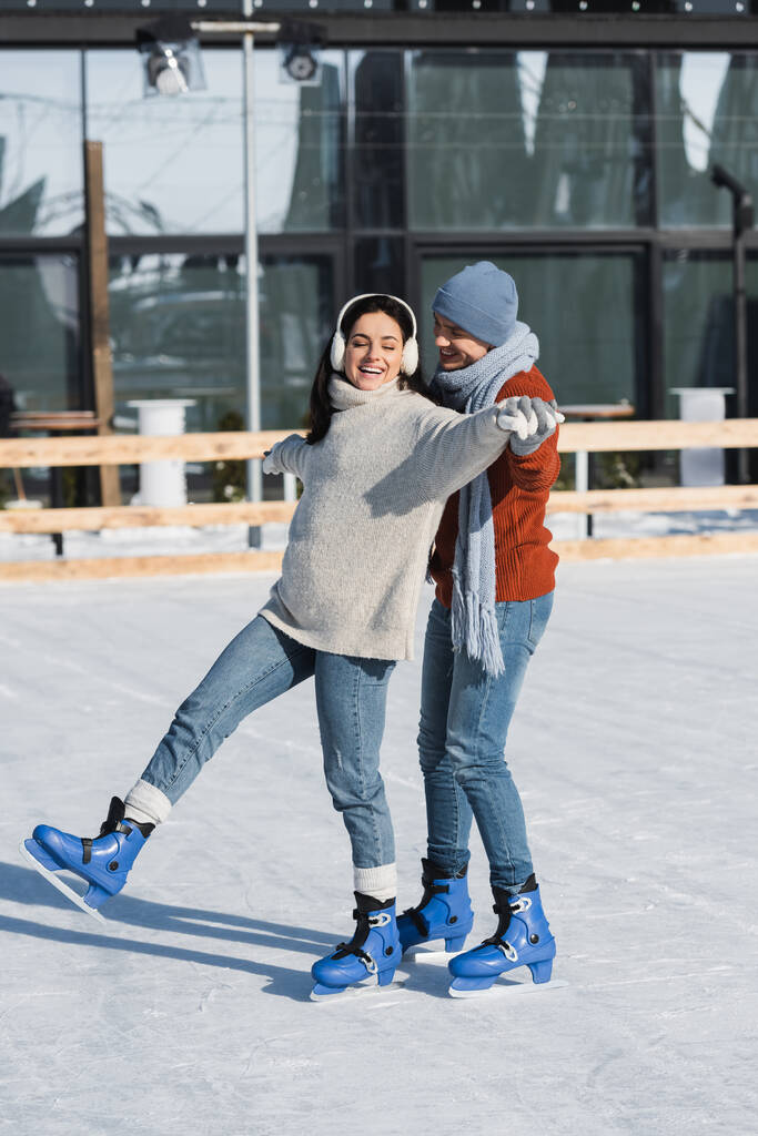 volle Länge der glücklichen Frau in Ohrenschützer Händchen halten mit fröhlichen Freund beim Schlittschuhlaufen auf der Eisbahn  - Foto, Bild