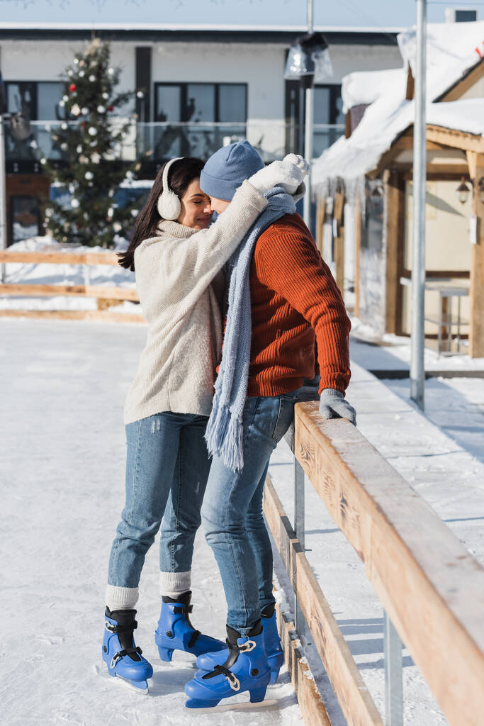 piena lunghezza di donna felice in manicotti per le orecchie abbracciando l'uomo in cappello invernale appoggiato sul bordo di legno sulla pista di pattinaggio  - Foto, immagini