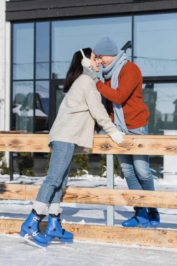 pełna długość szczęśliwa para uśmiechnięta podczas całowania w pobliżu drewnianej granicy na lodowisku  - Zdjęcie, obraz