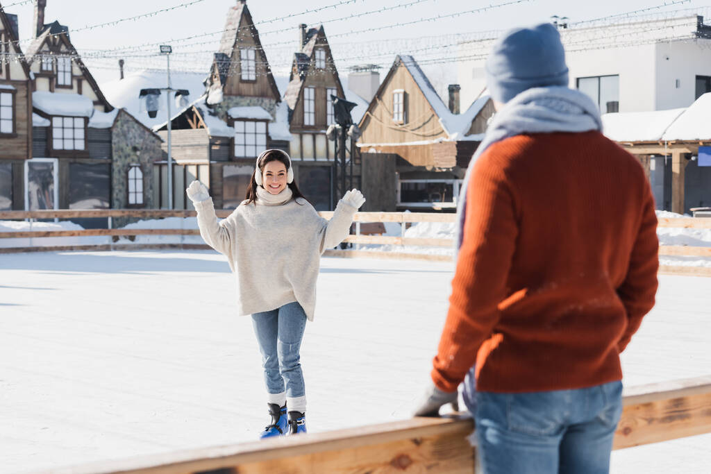 Buz patenli mutlu genç bir kadın ön planda bulanık erkek arkadaşına bakıyor. - Fotoğraf, Görsel