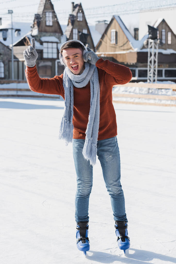 longitud completa de joven feliz en suéter y bufanda escuchando música mientras patina en pista de hielo  - Foto, Imagen