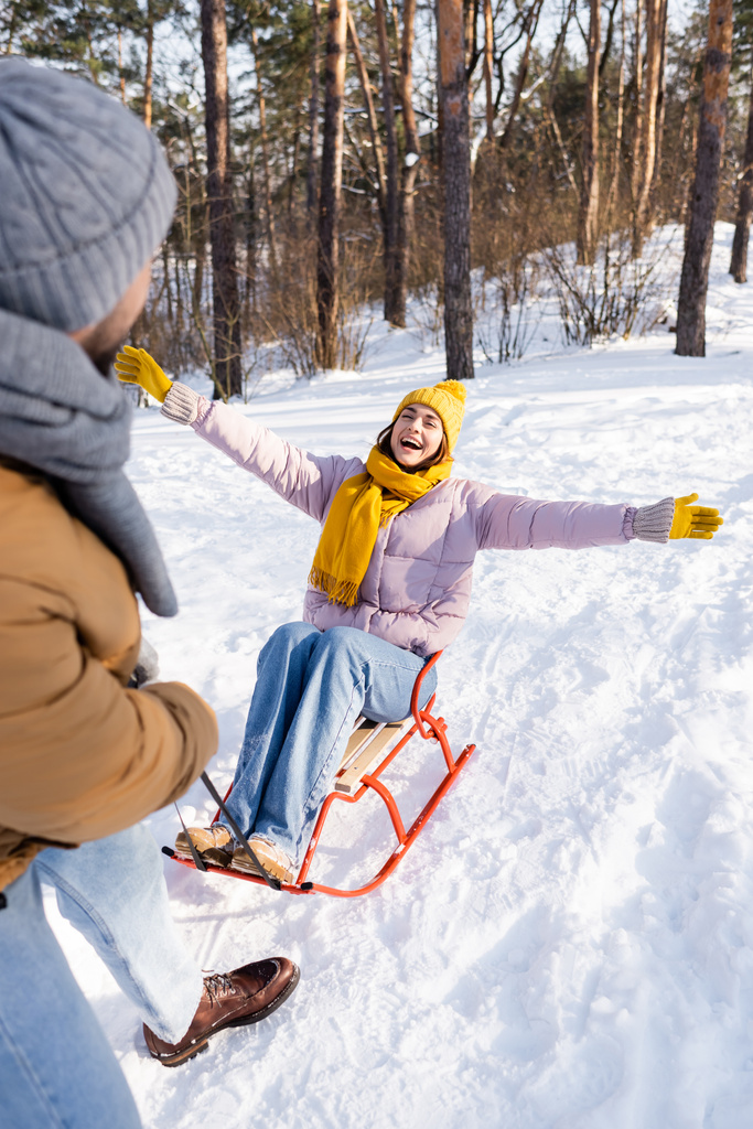 Cheerful woman sitting on sled near blurred boyfriend in winter park  - Photo, Image