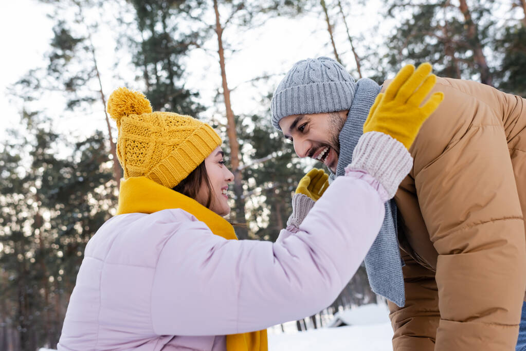 Вид сбоку на возбужденную пару в шарфах и шляпах в зимнем парке  - Фото, изображение