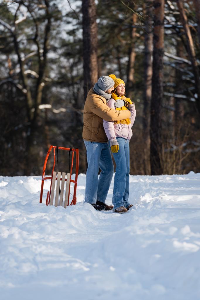 Giovane uomo che abbraccia sorridente ragazza vicino slitta nel parco invernale  - Foto, immagini