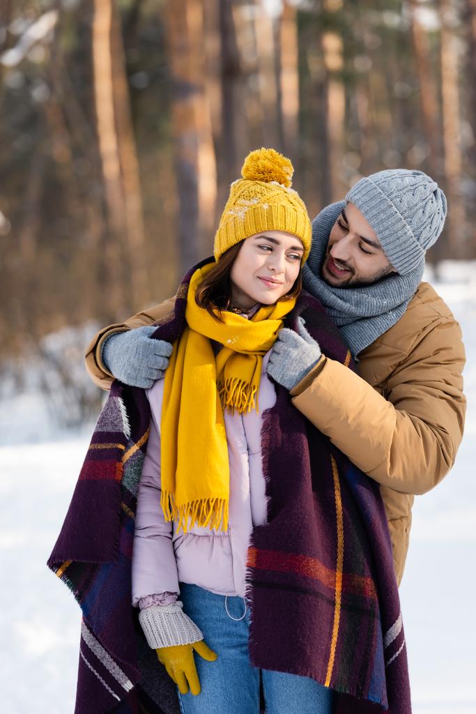 Улыбающийся мужчина в одеяле на своей девушке в зимнем парке  - Фото, изображение