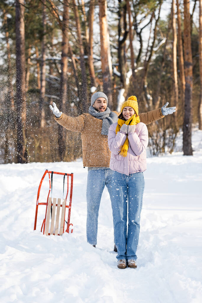 Wesoły człowiek rzuca śnieg w pobliżu dziewczyny z zamkniętymi oczami i sanki w parku zimowym  - Zdjęcie, obraz