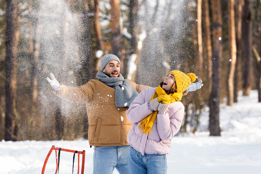 Podekscytowana kobieta stojąca obok chłopaka rzucająca śnieg i sanie w parku  - Zdjęcie, obraz