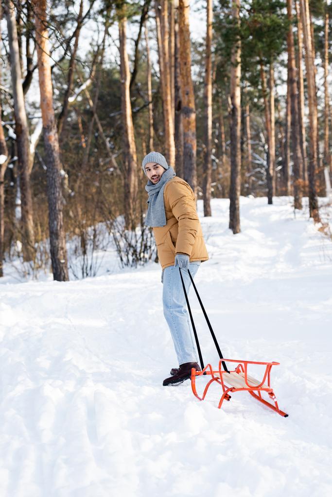 Людина в капелюсі і шарфі посміхається на камеру, тягнучи санчата в зимовому парку
  - Фото, зображення