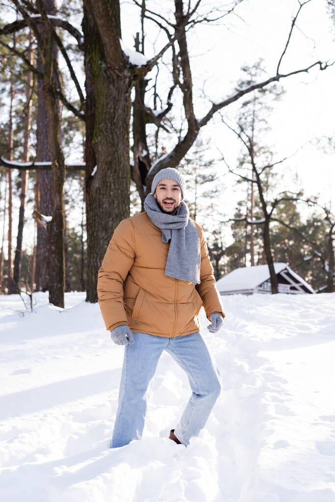 Fröhlicher Mann mit warmem Schal und Hut steht im verschneiten Park  - Foto, Bild