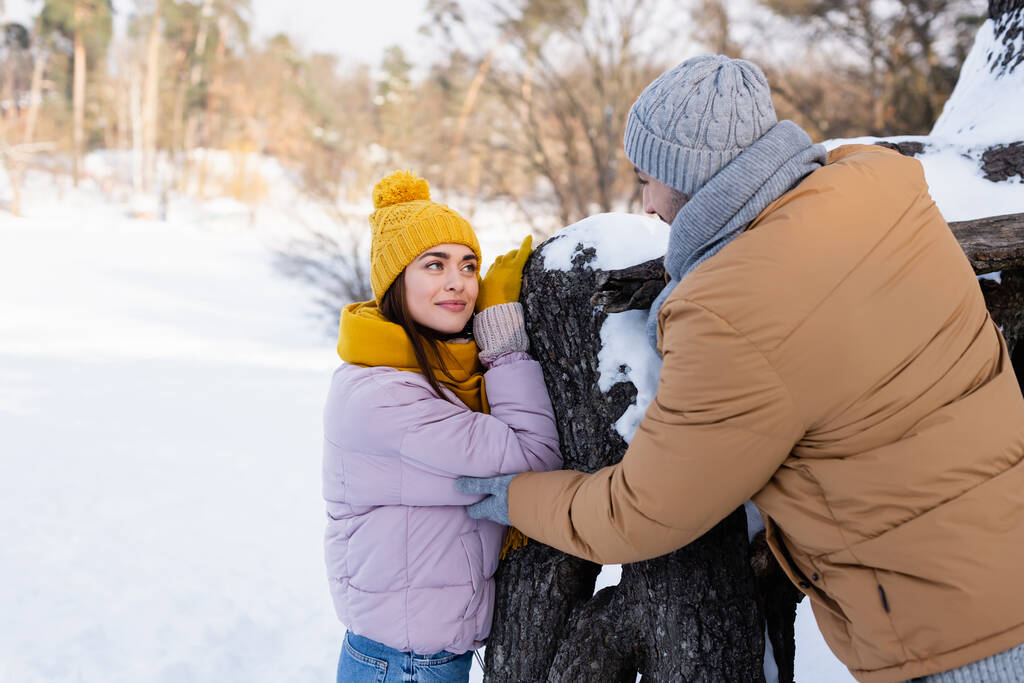 Молодая женщина смотрит на парня в зимнем наряде рядом с деревом в парке  - Фото, изображение
