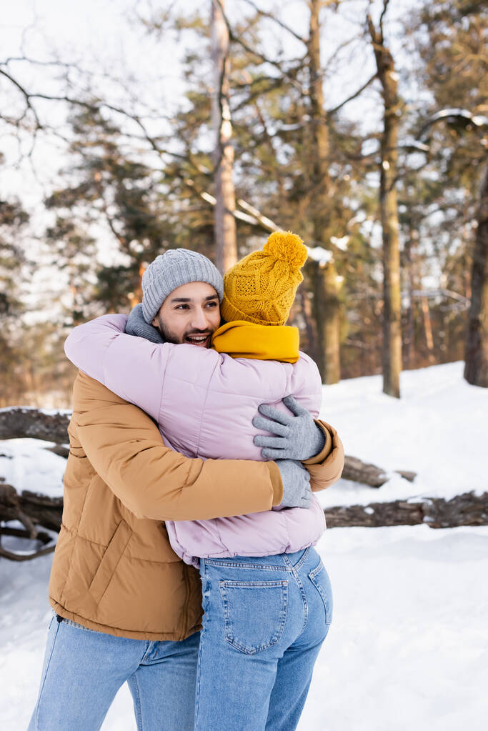 Улыбающийся мужчина обнимает девушку в зимнем наряде в парке днем  - Фото, изображение