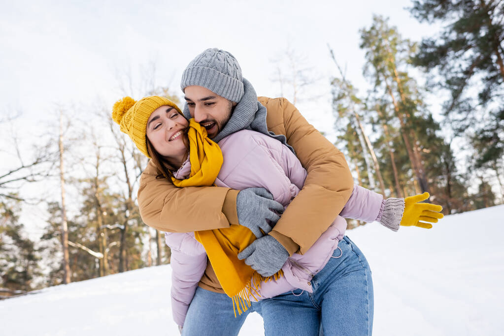 vzrušený muž v pleteném klobouku objímající přítelkyni se zavřenýma očima v parku se sněhem  - Fotografie, Obrázek