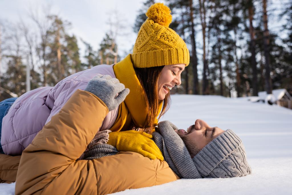 Örgü şapkalı olumlu bir kadının parkta karda erkek arkadaşının üzerine uzanması.  - Fotoğraf, Görsel