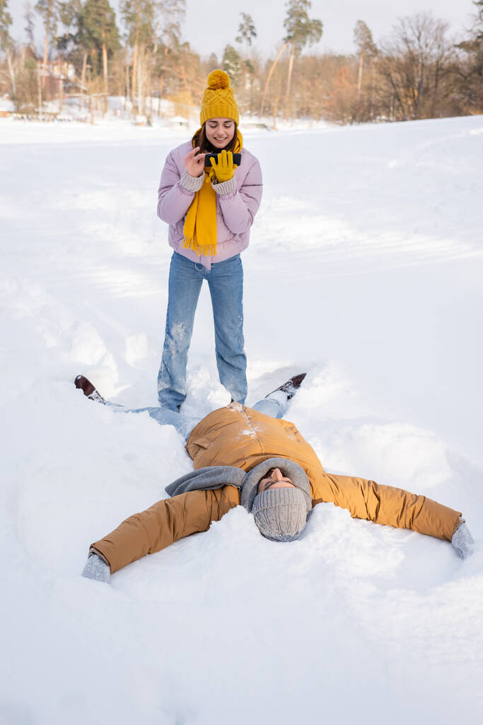 glimlachende vrouw het nemen van foto terwijl vriendje het maken van sneeuw engel in park  - Foto, afbeelding