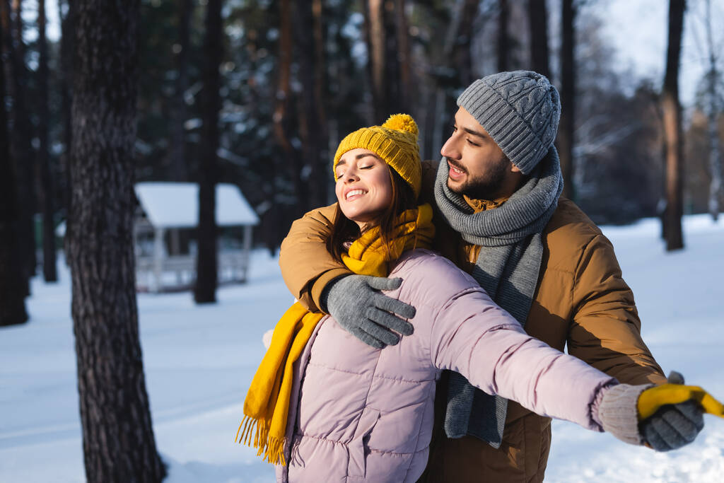 Мужчина обнимает и держит за руку счастливую девушку с закрытыми глазами в зимнем парке  - Фото, изображение