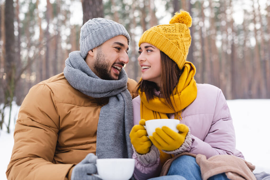 Счастливый человек в вязаной шляпе держит чашку возле подружки в парке зимой - Фото, изображение