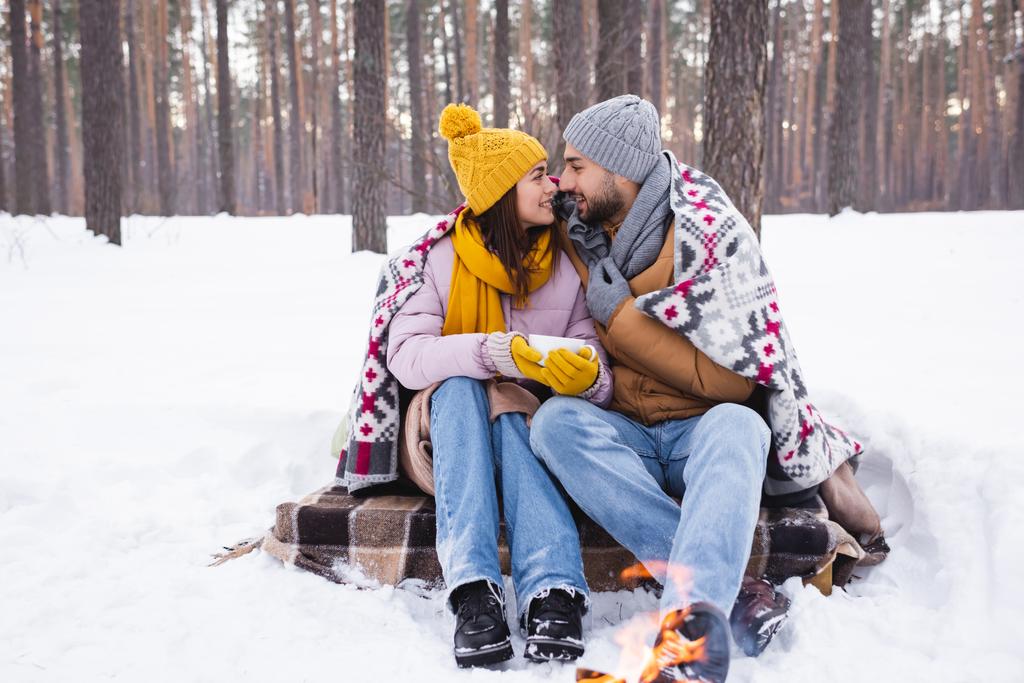 Vista lateral de pareja positiva con taza y manta mirándose cerca de hoguera borrosa en el parque de invierno  - Foto, Imagen