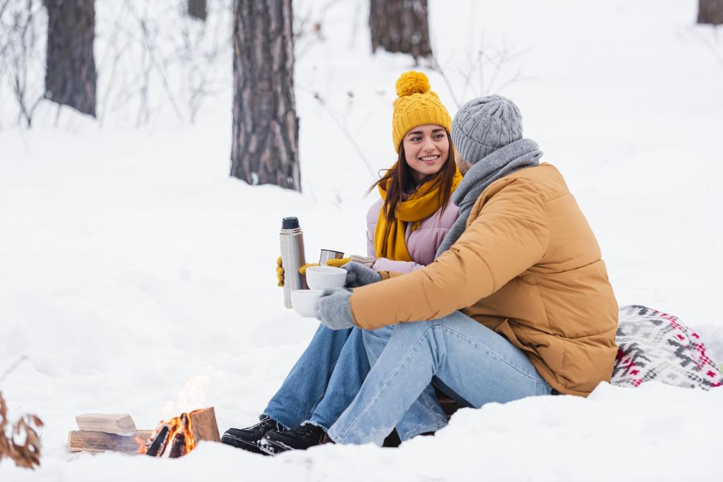 Мужчина держит чашки возле улыбающейся подруги с термосом и костром в снежном парке  - Фото, изображение