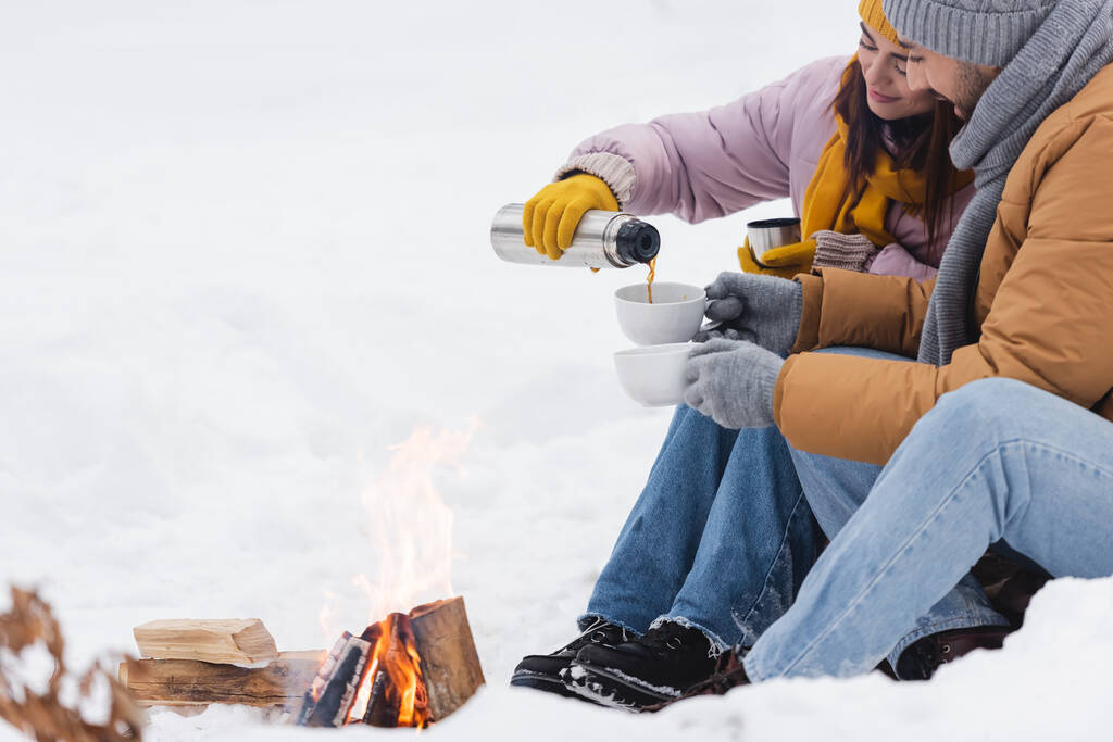 Žena nalévání kávy z termosky v blízkosti přítele a táborák v zimním parku  - Fotografie, Obrázek