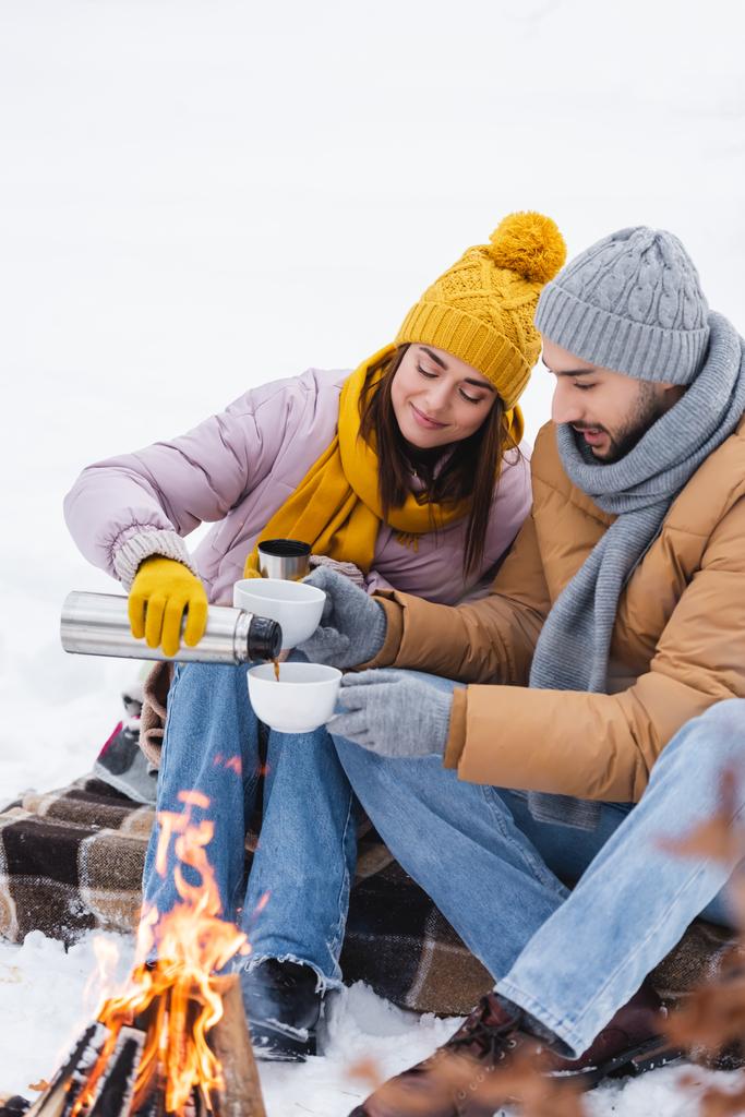 Bir adam, kız arkadaşının yanında termoslu bardak tutuyor ve kış parkında şenlik ateşini bulanıklaştırıyor.  - Fotoğraf, Görsel
