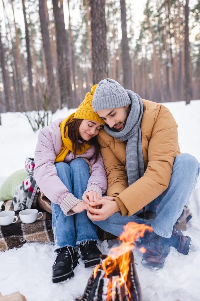 Νεαρό ζευγάρι σε χειμερινό ντύσιμο προειδοποιητικά χέρια κοντά σε φωτιά στο πάρκο  - Φωτογραφία, εικόνα