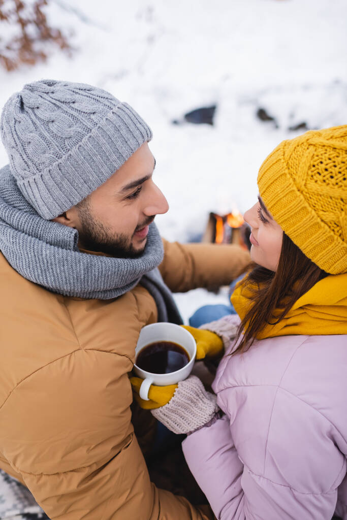 Вид женщины в вязаной шляпе с чашкой кофе рядом с улыбающимся парнем в зимнем парке  - Фото, изображение