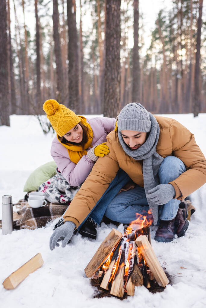 Glimlachende vrouw knuffelende vriend in de buurt van bekers, thermoskan en kampvuur in winterpark  - Foto, afbeelding