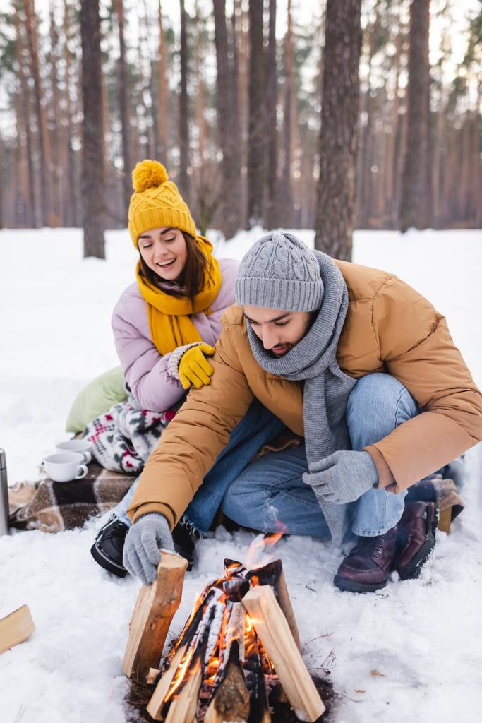 Femme souriante regardant petit ami avec des bûches de bois près de feu de joie dans le parc d'hiver  - Photo, image