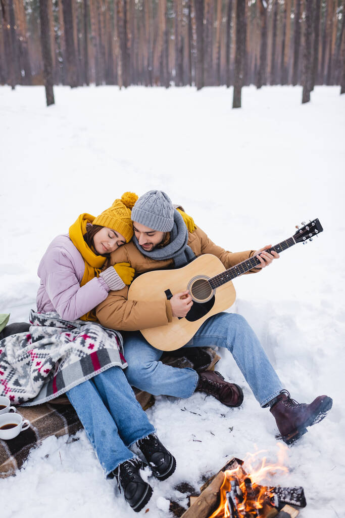 Kız arkadaşının yanında akustik gitar çalan gülümseyen adam ve kış parkında şenlik ateşi.  - Fotoğraf, Görsel