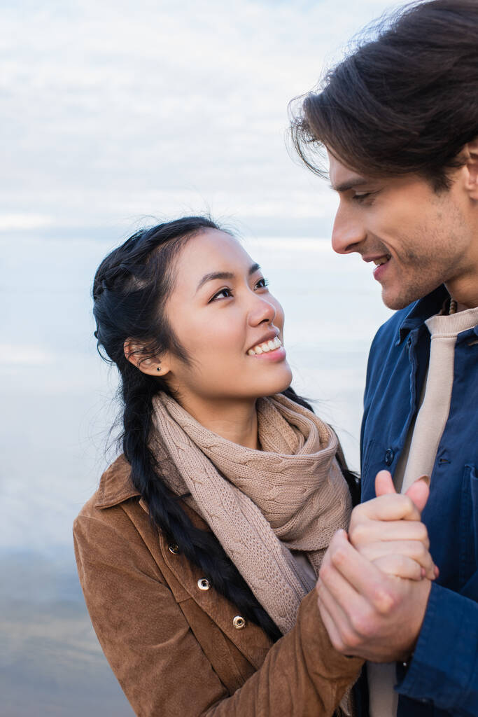 Sorridente coppia multietnica che si tiene per mano mentre si guarda vicino al lago  - Foto, immagini