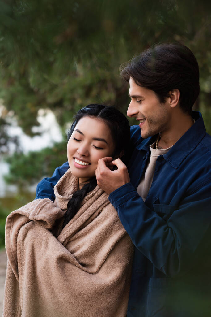 Усміхнений чоловік торкається щоки азіатської дівчини в ковдрі на відкритому повітрі
  - Фото, зображення