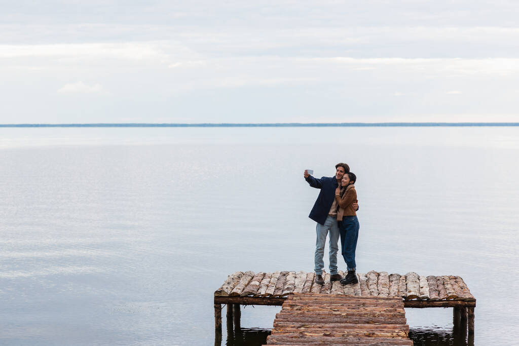 Boldog többnemzetiségű pár szelfit készít mobilon a mólón, közel a tengerhez  - Fotó, kép