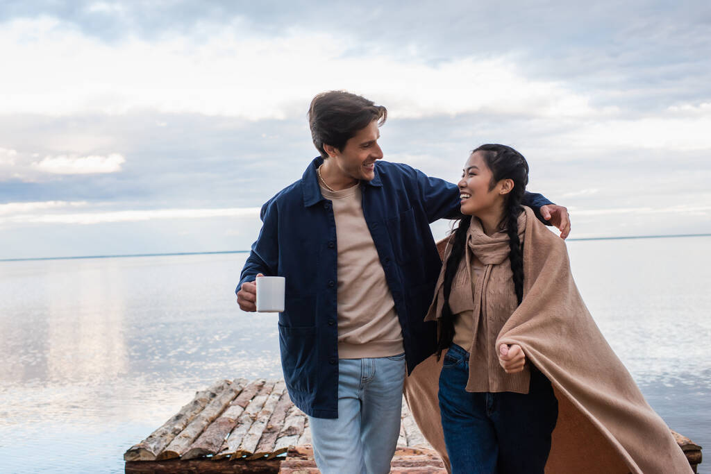 Lächelnder Mann mit Tasse umarmt asiatische Freundin in Decke auf Pier  - Foto, Bild