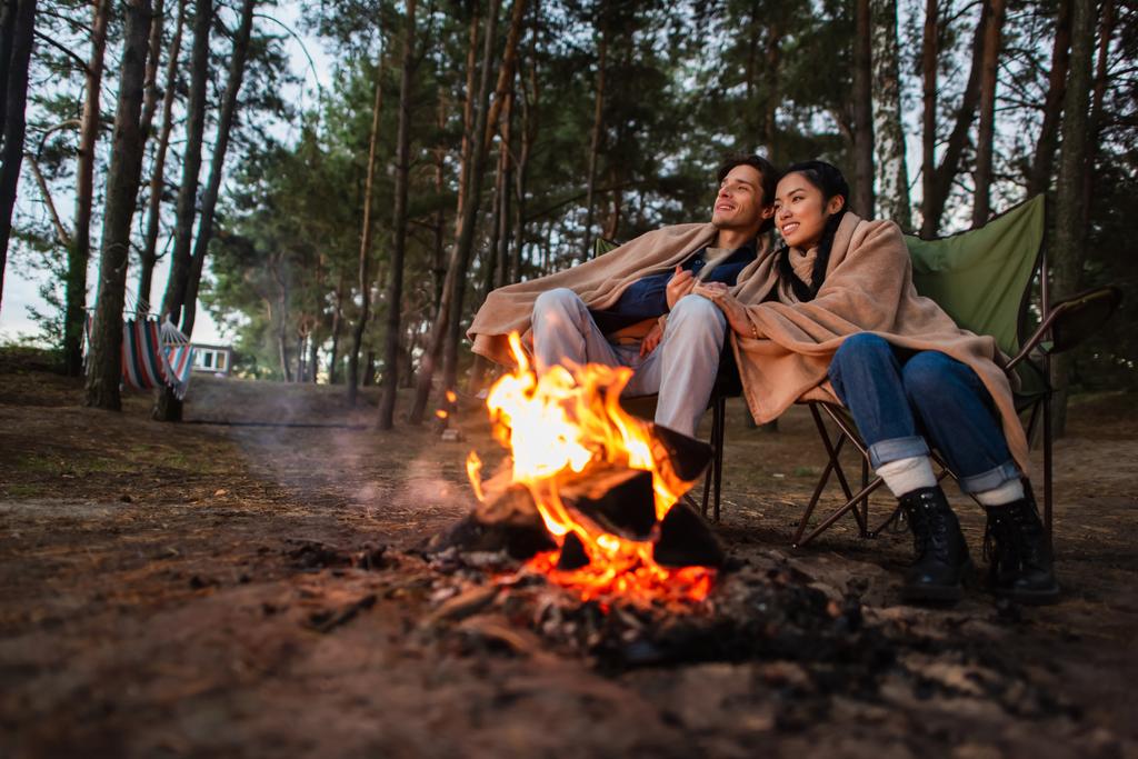 Jeune couple multiethnique dans des couvertures assis près du feu de camp flou  - Photo, image