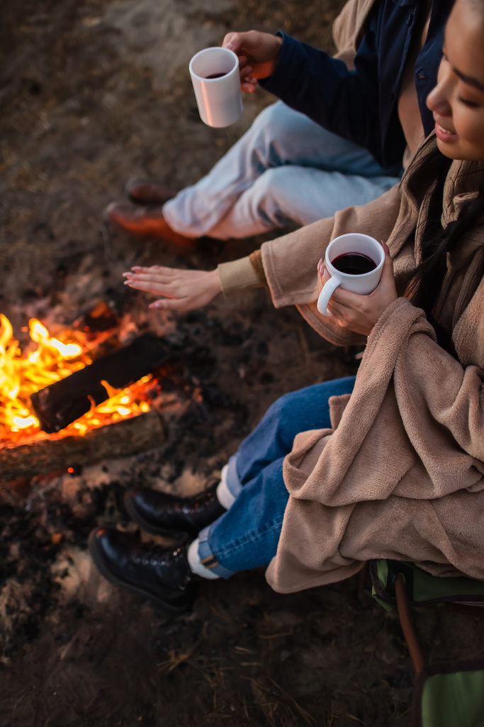 High angle view of smiling asian woman holding cup near boyfriend and campfire  - Photo, Image