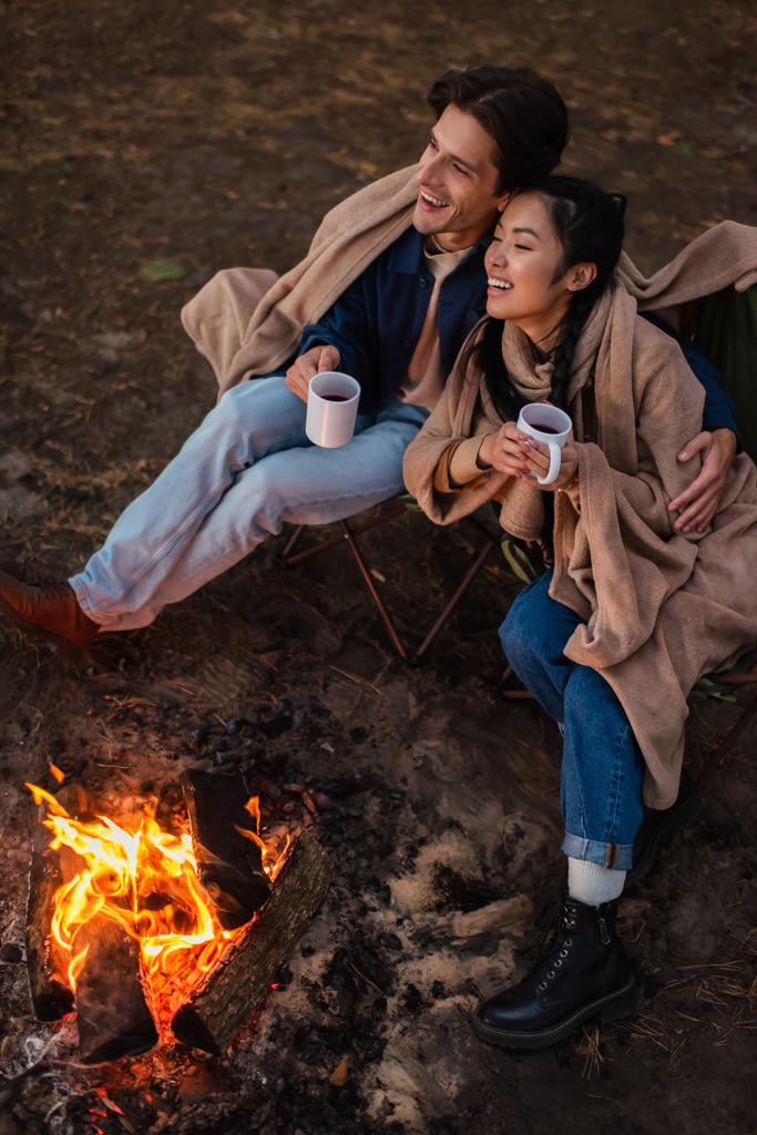 Uśmiechnięta para międzyrasowa z filiżankami przytulającymi się przy ognisku podczas weekendu  - Zdjęcie, obraz